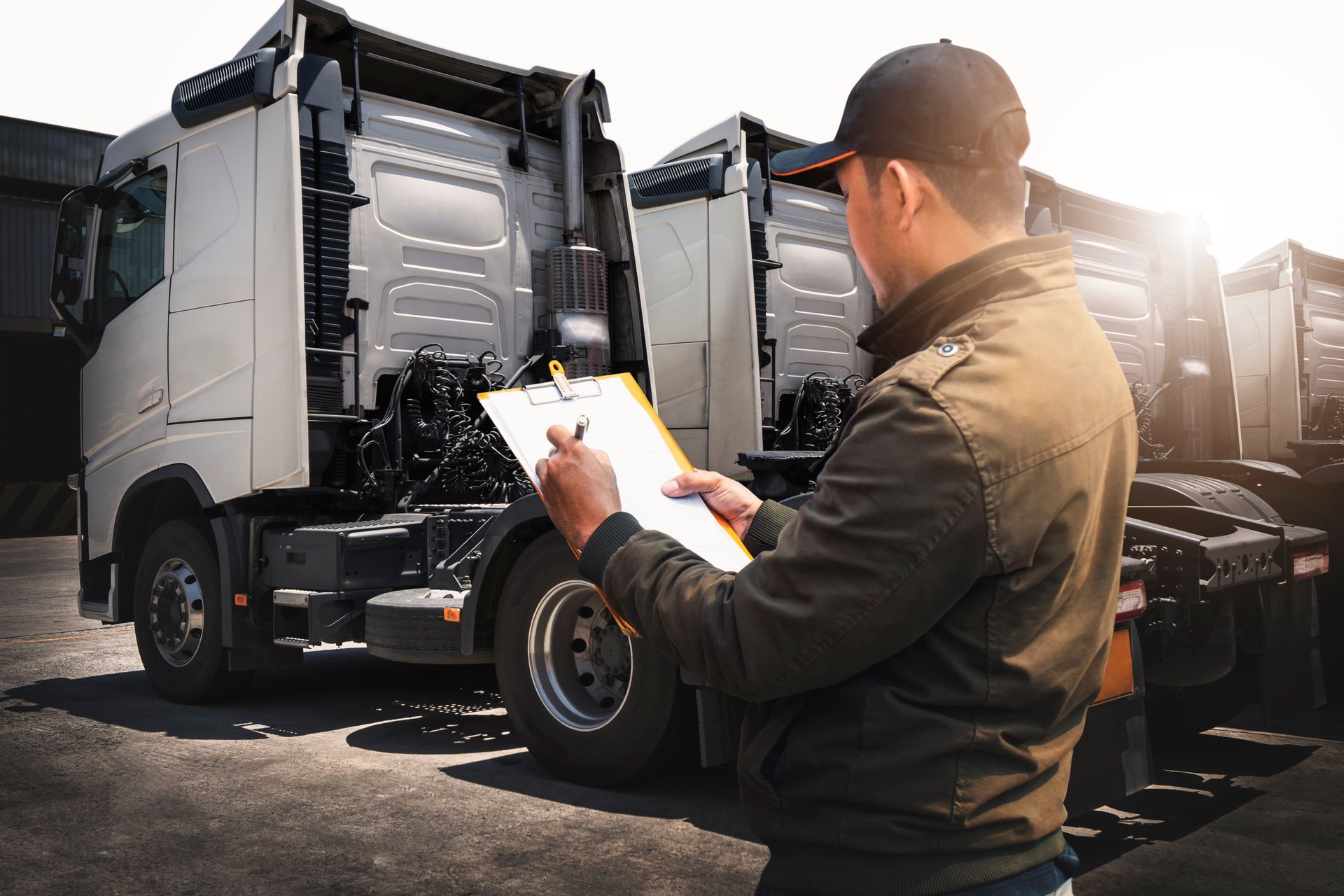 Man in cap and jacket, writing on a clipboard while inspecting trucks in a lot.