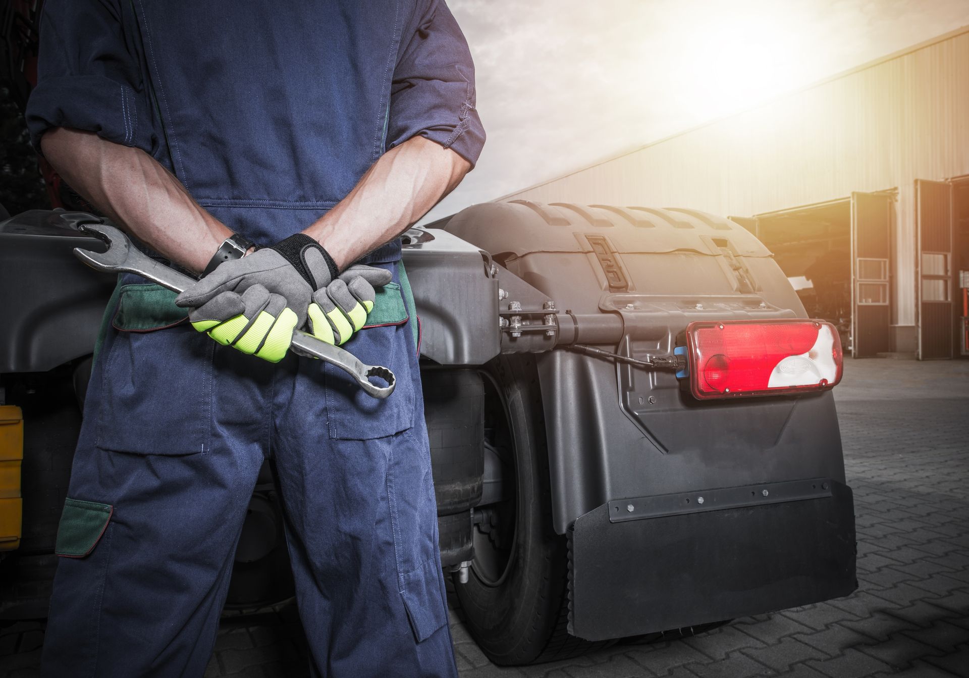Mechanic in blue overalls holding a wrench in front of a truck, near a sunny garage.