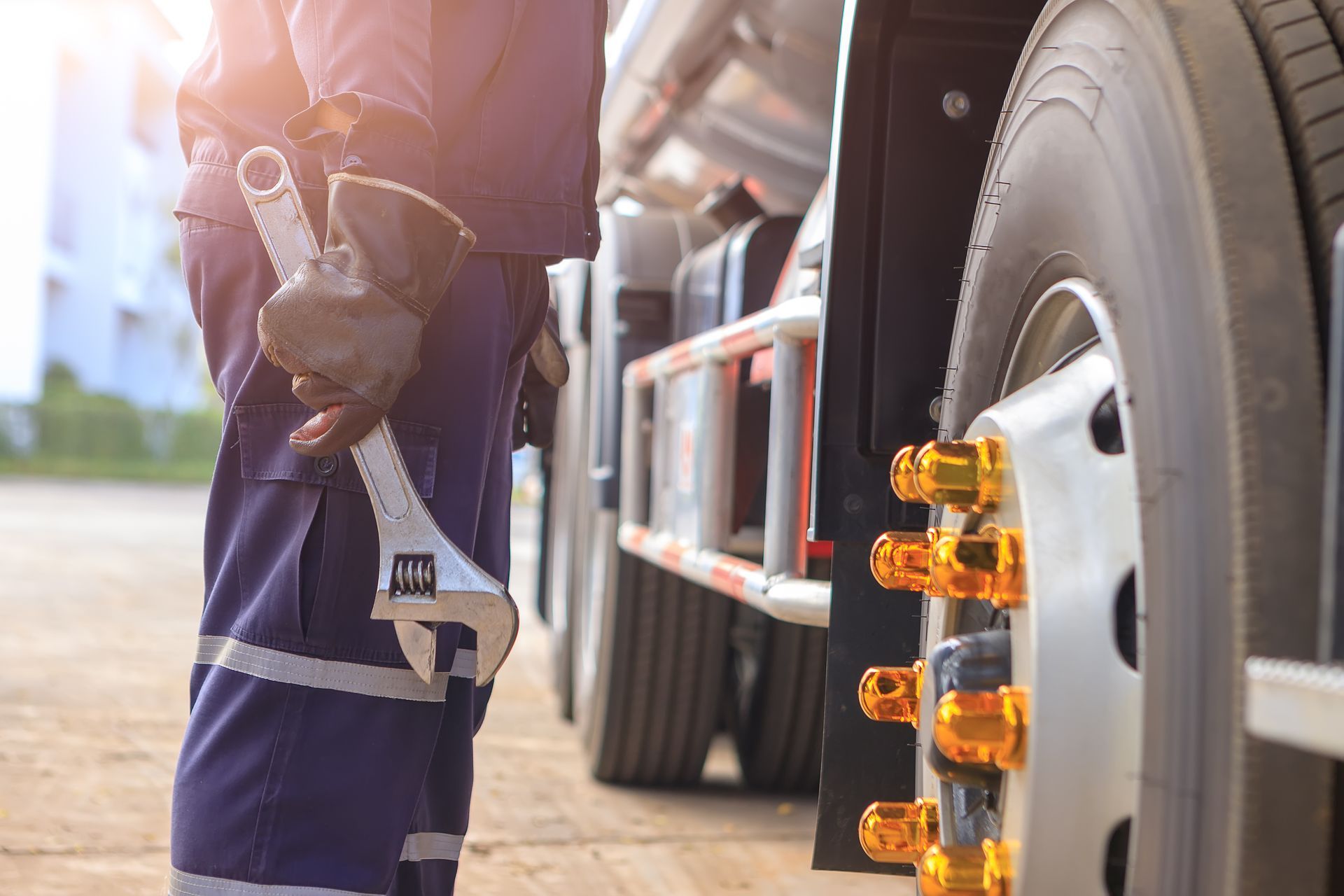 Mechanic holding wrench next to truck wheel. Blue overalls, safety gloves, outdoor setting.