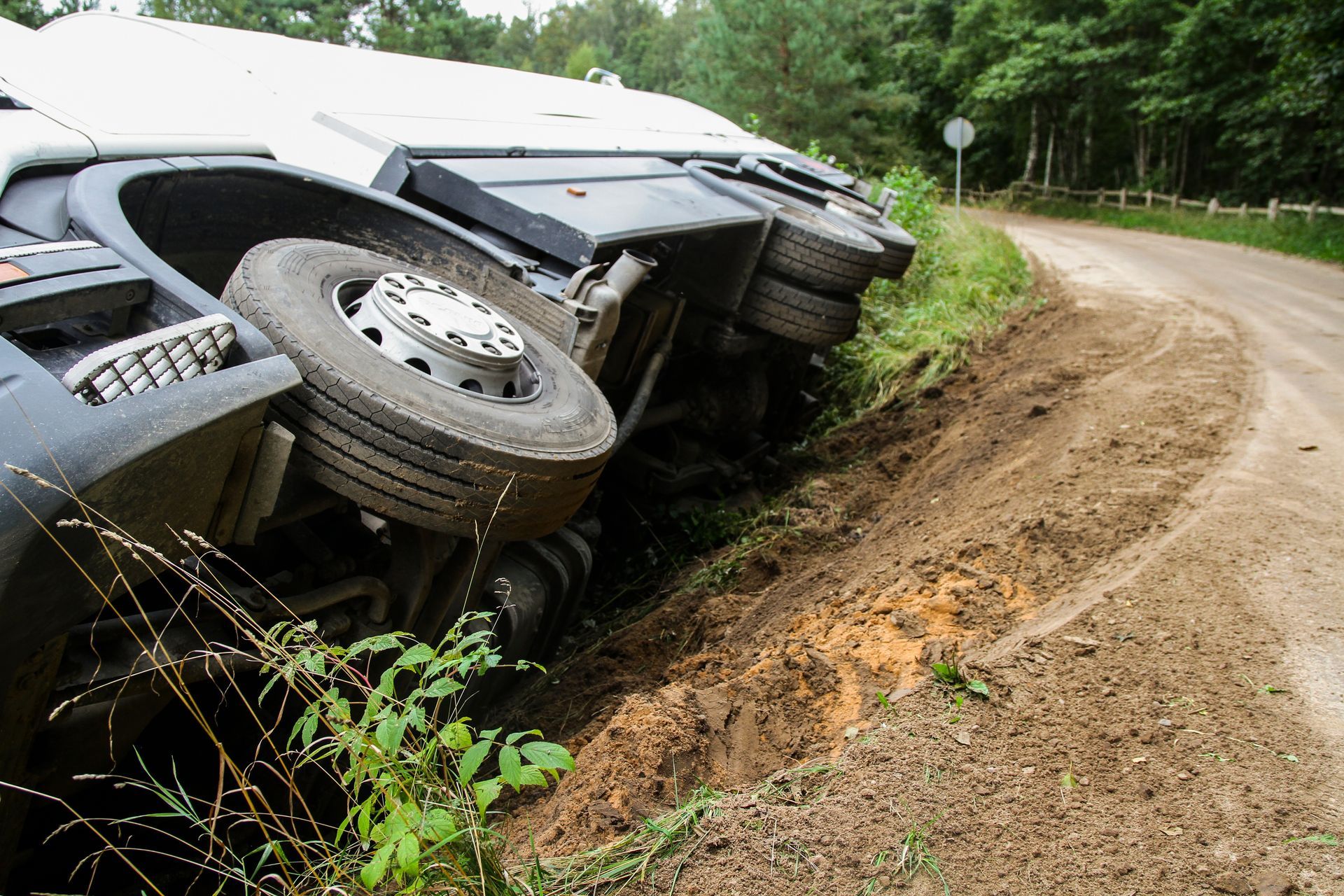 Truck overturned off the side of a dirt road, wheels visible, surrounded by dirt and vegetation.