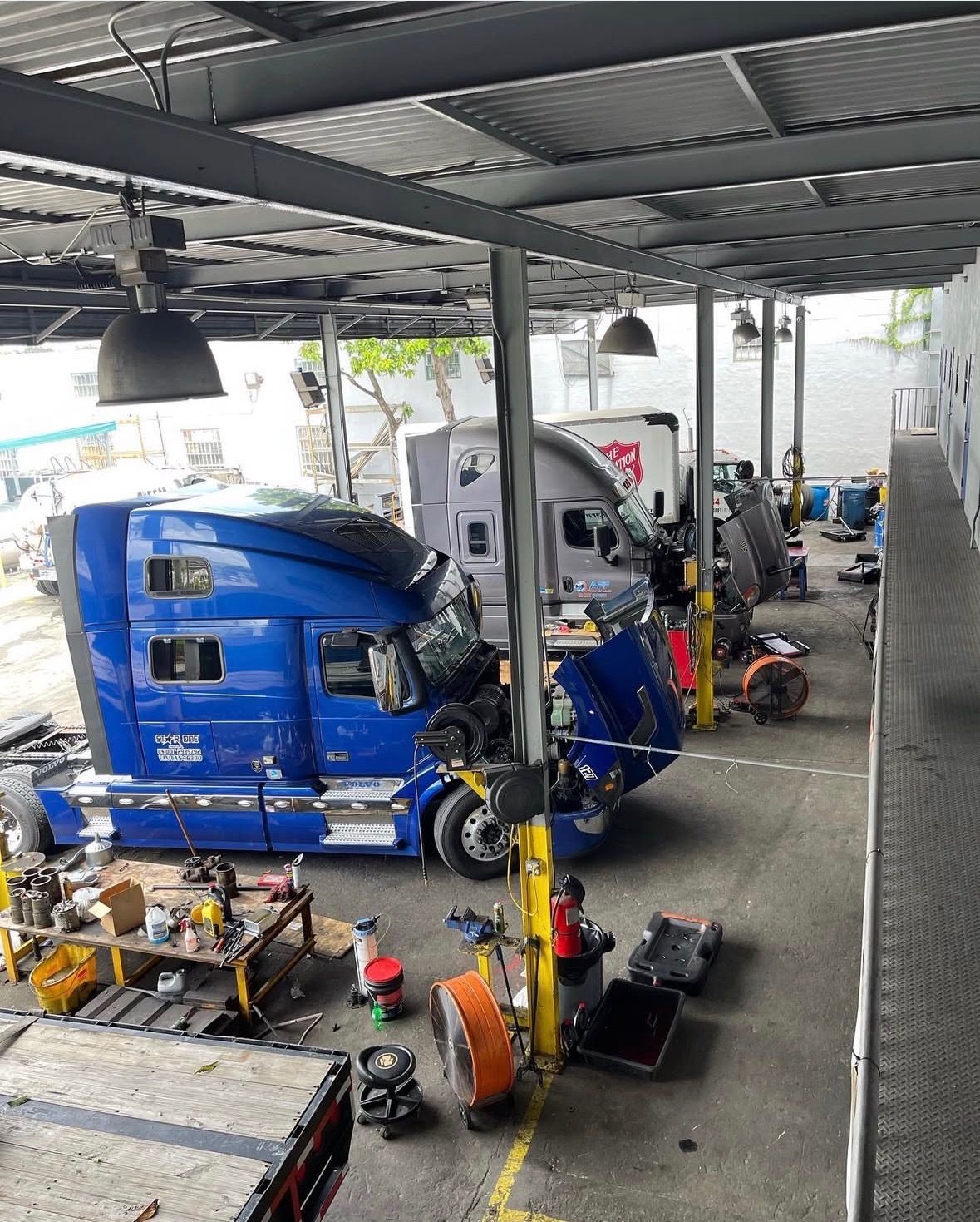 Trucks in a repair bay, some disassembled. Blue and gray semi-trucks under a metal roof, tools scattered.