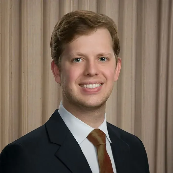 Smiling man in a suit and tie, against a light brown striped background.