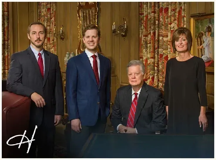 Four formally dressed people in a room with ornate decor; two men standing, one seated, and one woman standing.