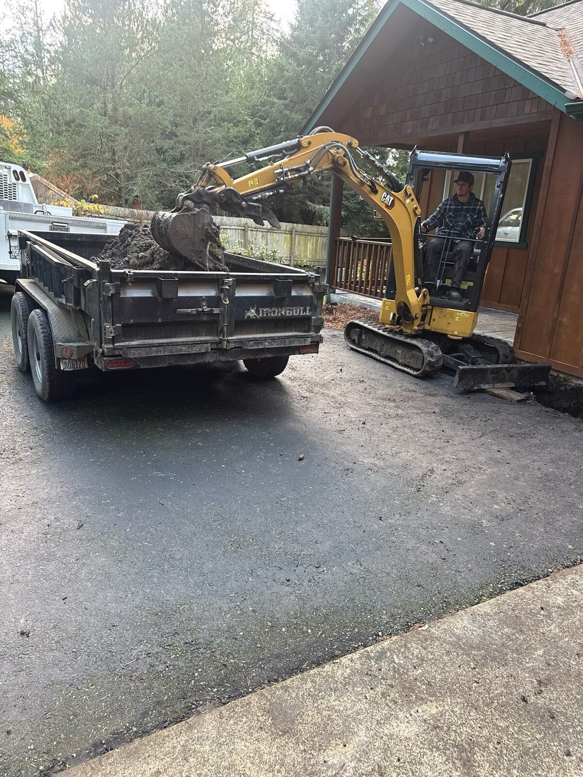 A yellow excavator dumps soil into a black dump trailer parked on a paved driveway next to a wooden building.