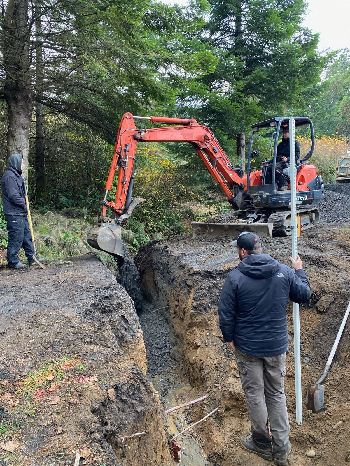 An orange excavator digs a trench in a wooded area, while two people stand nearby observing the work.