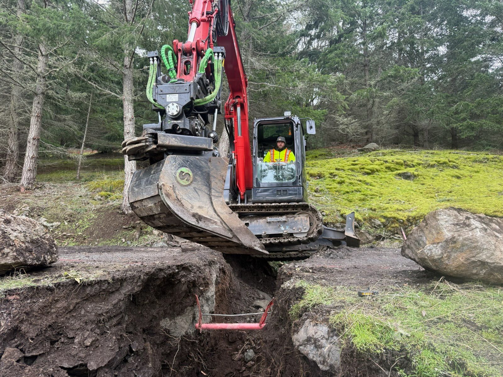A red excavator with a hydraulic thumb attachment digs a trench in a grassy, wooded area.