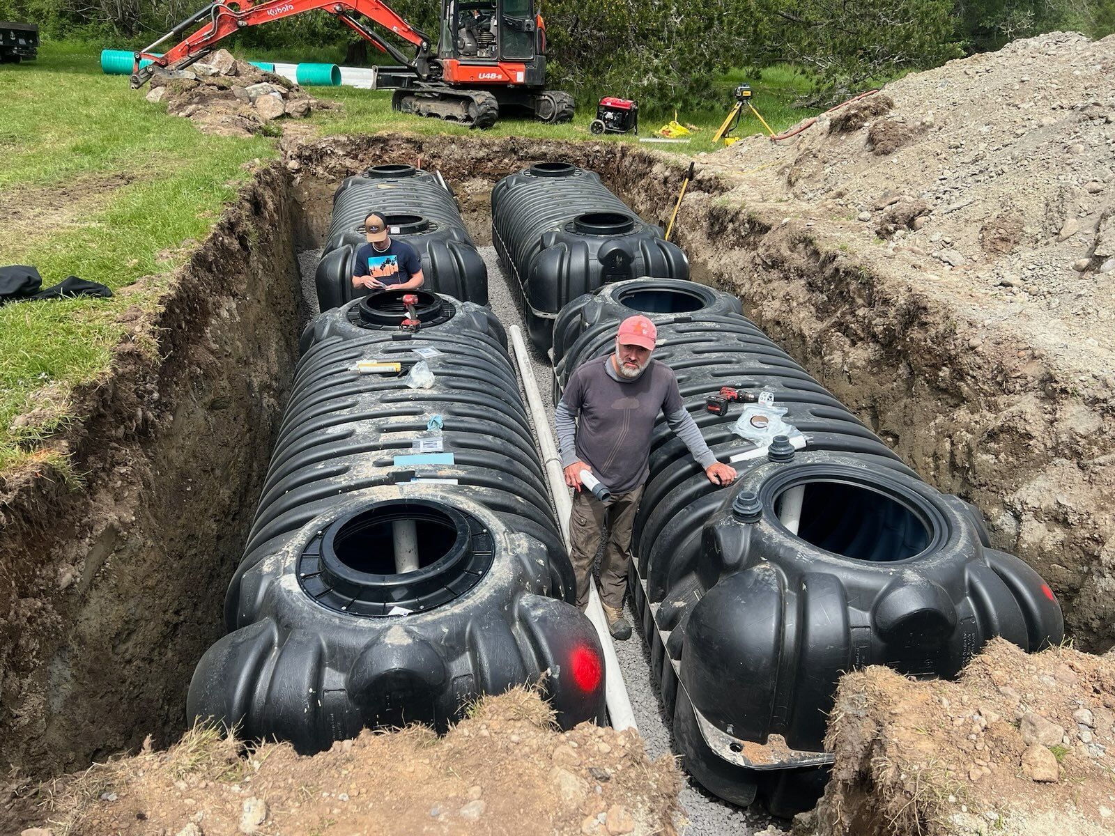Two people stand in a deep trench installing two large, black, ribbed plastic septic tanks in an outdoor, grassy area.