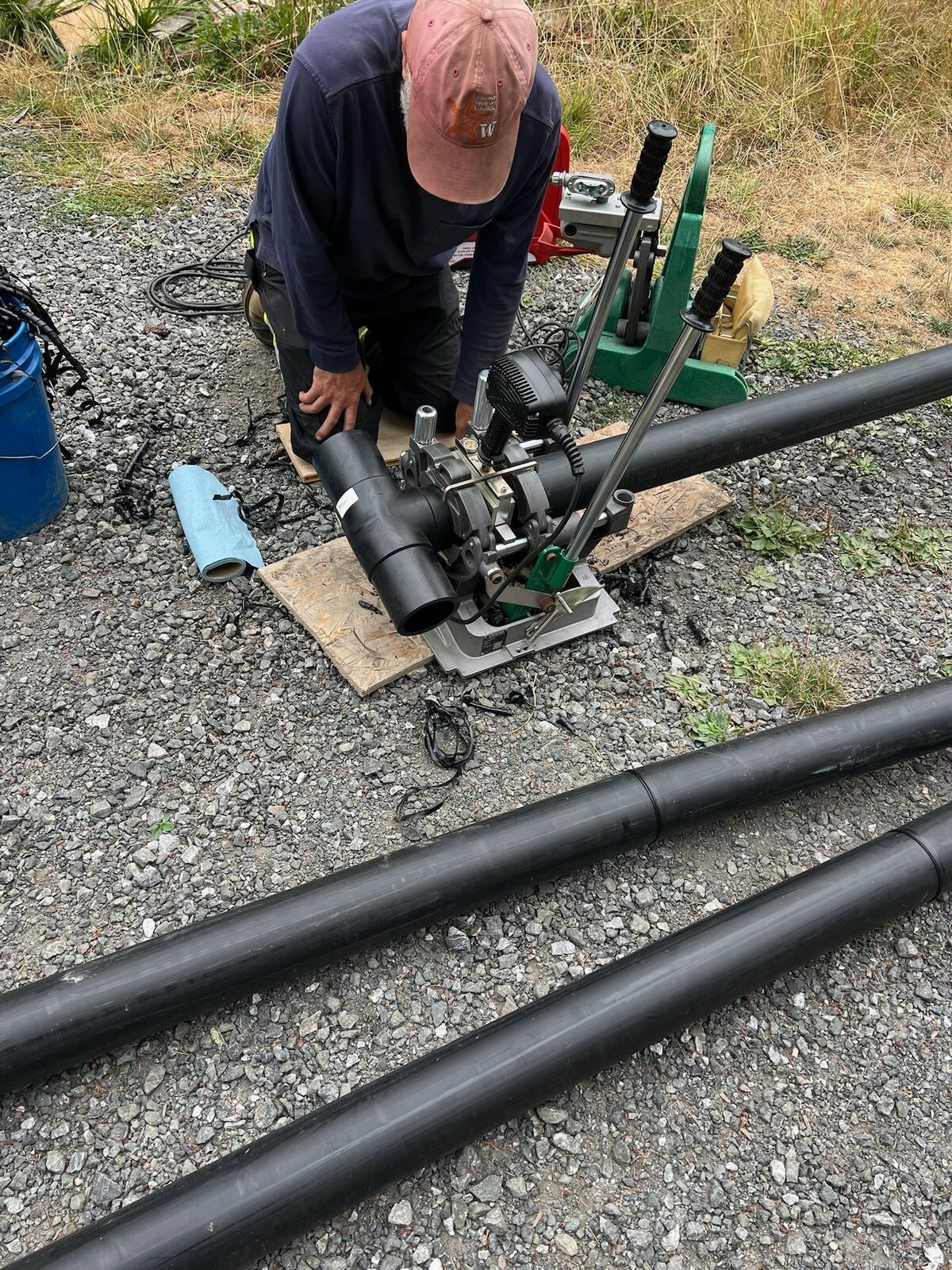 A person in a pink cap and dark clothing kneeling on gravel to operate a pipe fusion tool on black plastic pipes.