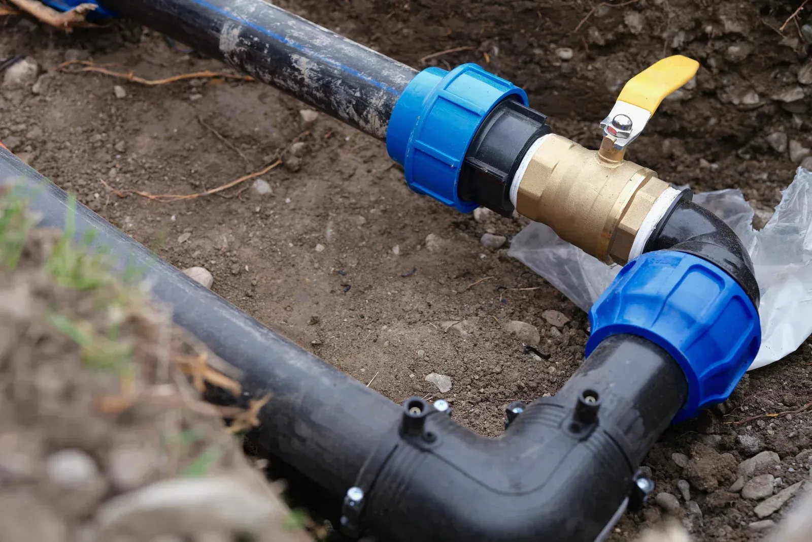 A black irrigation pipe with blue fittings and a yellow-handled brass ball valve installed in a dirt trench.