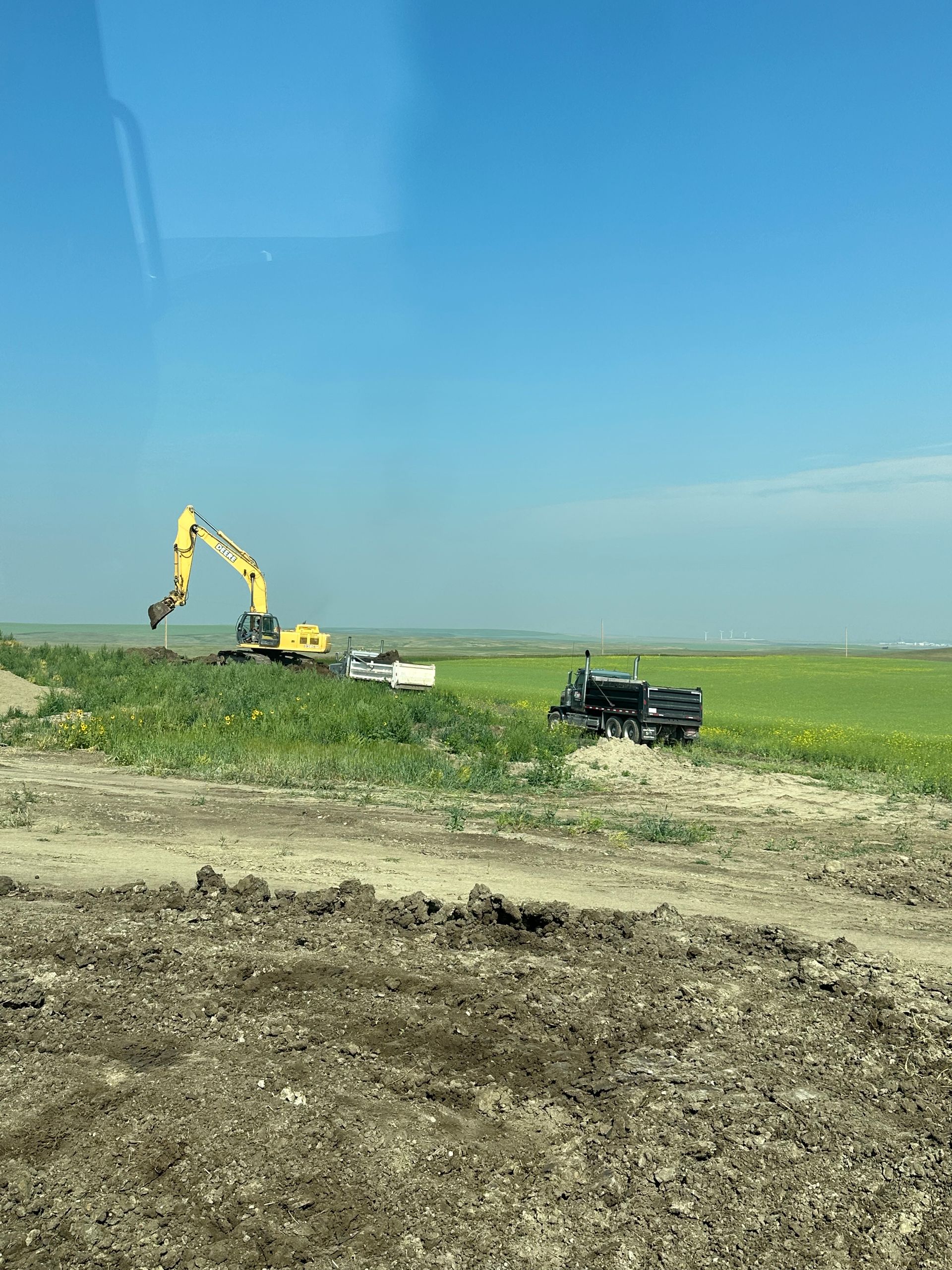 A truck is parked in the middle of a dirt field with a yellow excavator in the background.
