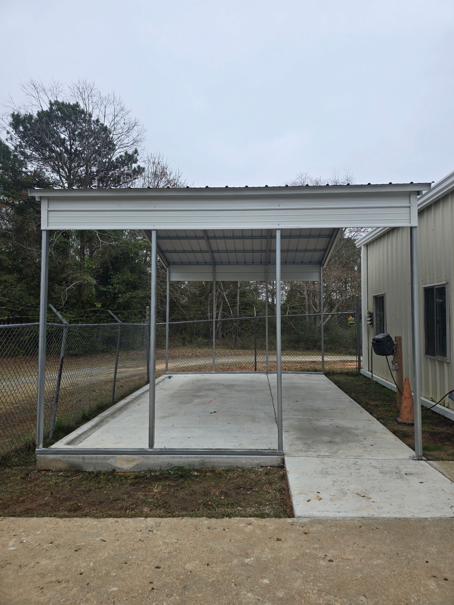 Metal carport with concrete base and adjoining walkway, adjacent to a building. Overcast sky.
