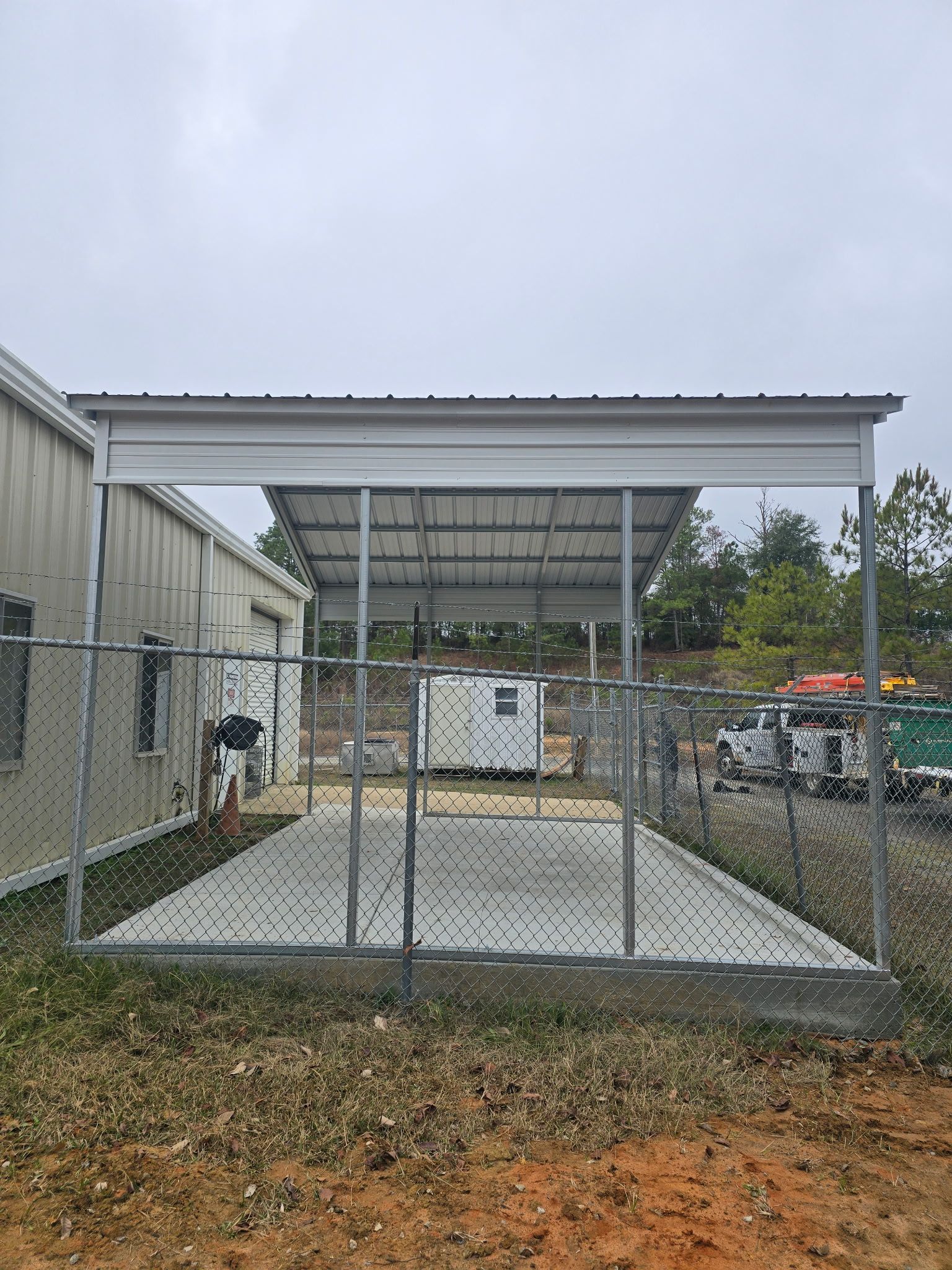 A metal carport with a chain-link fence on a concrete pad. A white building is to the left. Overcast day.
