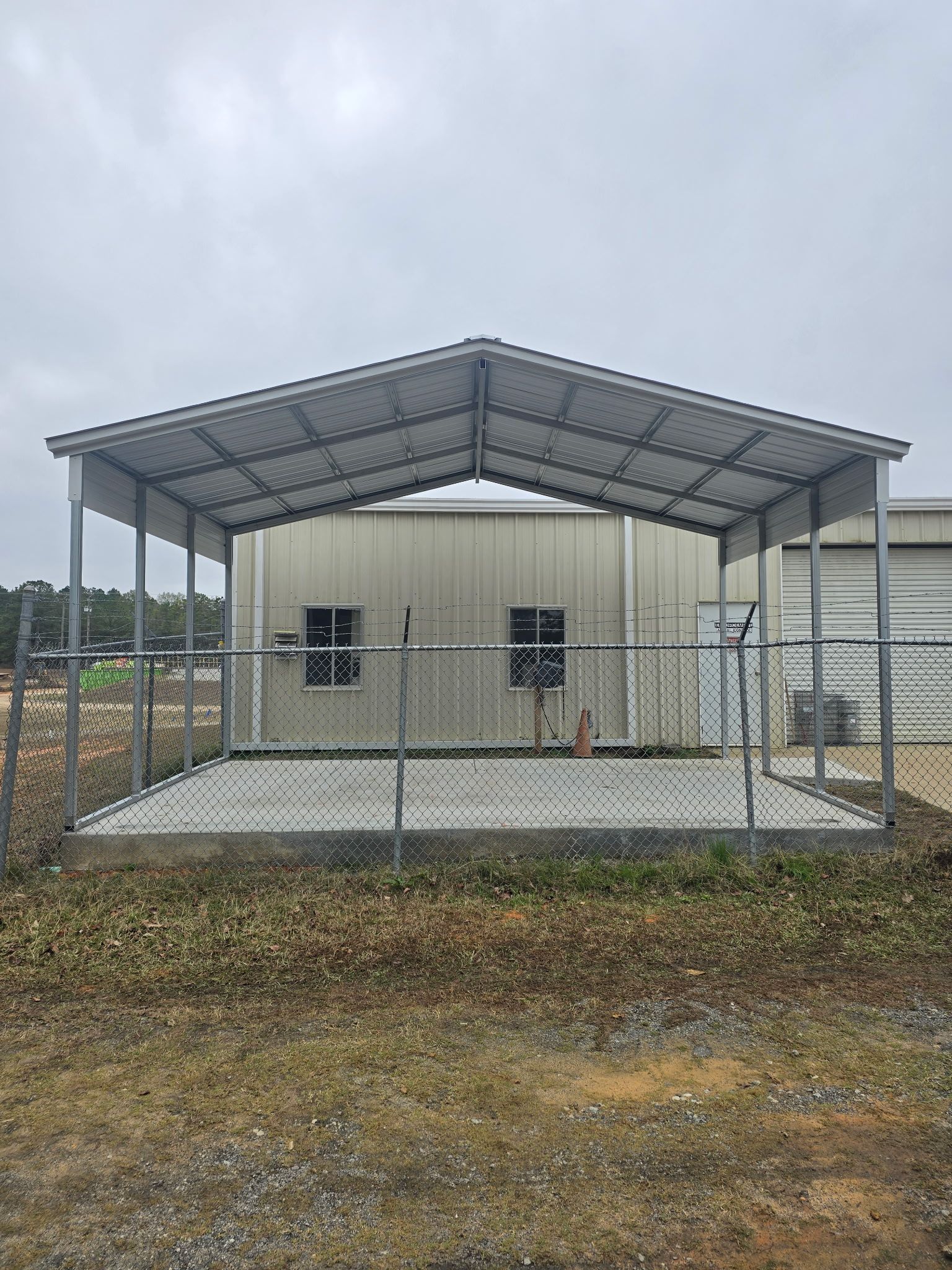 Metal shed with a chain-link fence in front of a tan industrial building, overcast sky.