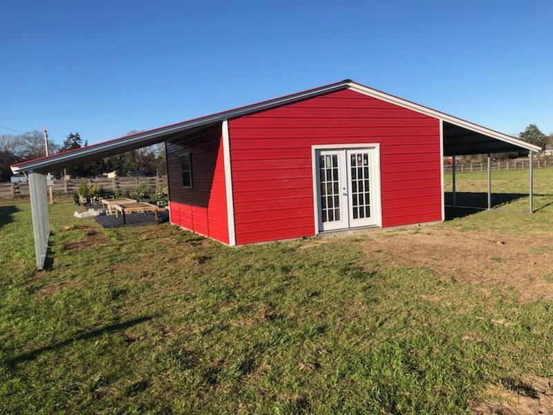 A red barn with a canopy over it is sitting in the middle of a grassy field.