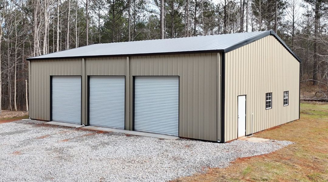 A large metal garage with three garage doors is sitting on top of a gravel driveway.