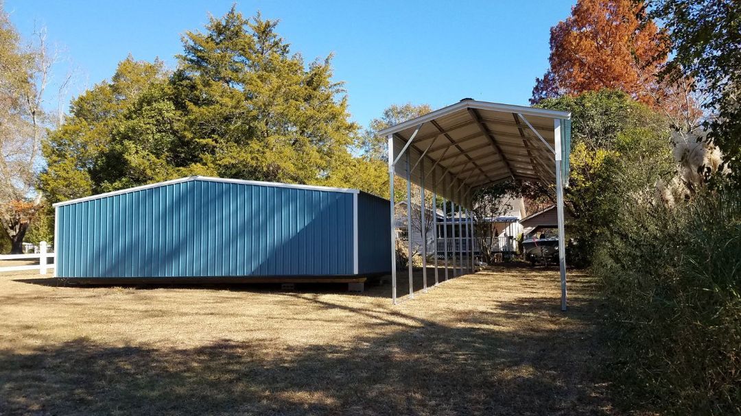 A blue trailer is parked under a carport in a field.
