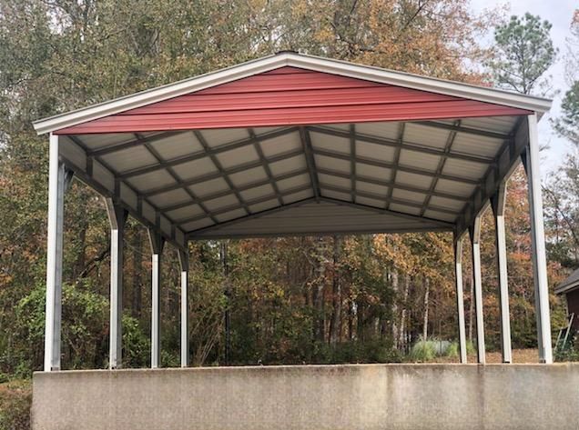 A red and white metal carport is sitting on top of a concrete foundation in the middle of a forest.