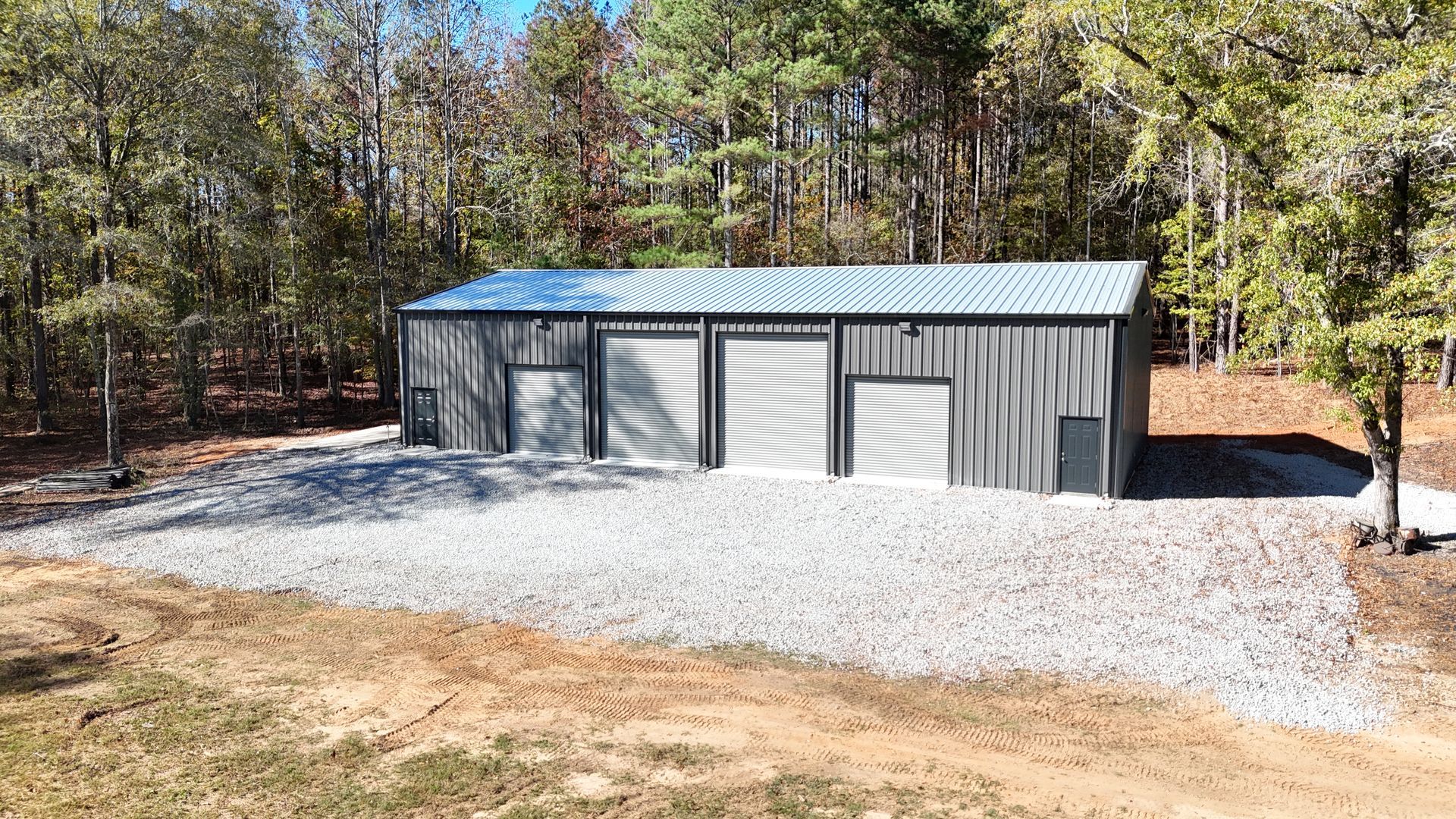 A large metal building is sitting on top of a gravel lot in the middle of a forest.