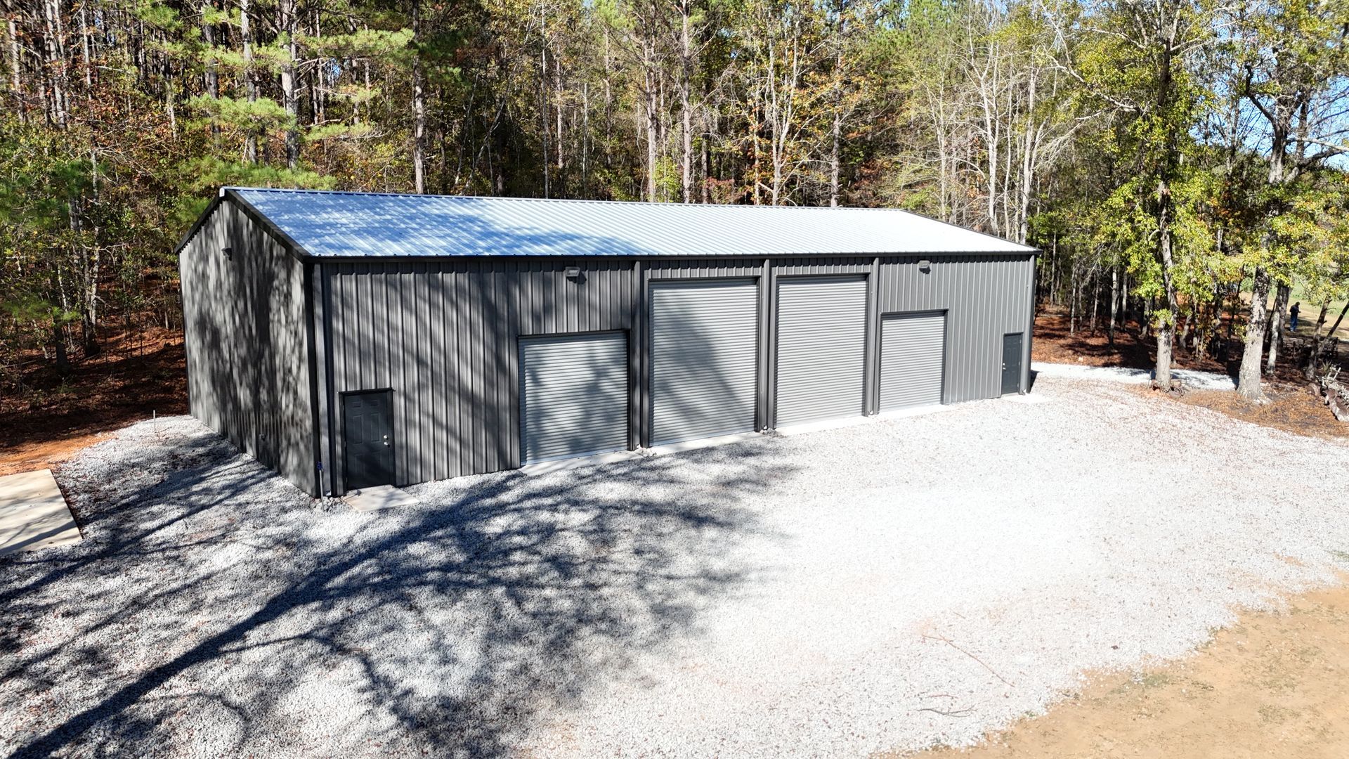 A large metal building is sitting on top of a gravel road in the middle of a forest.
