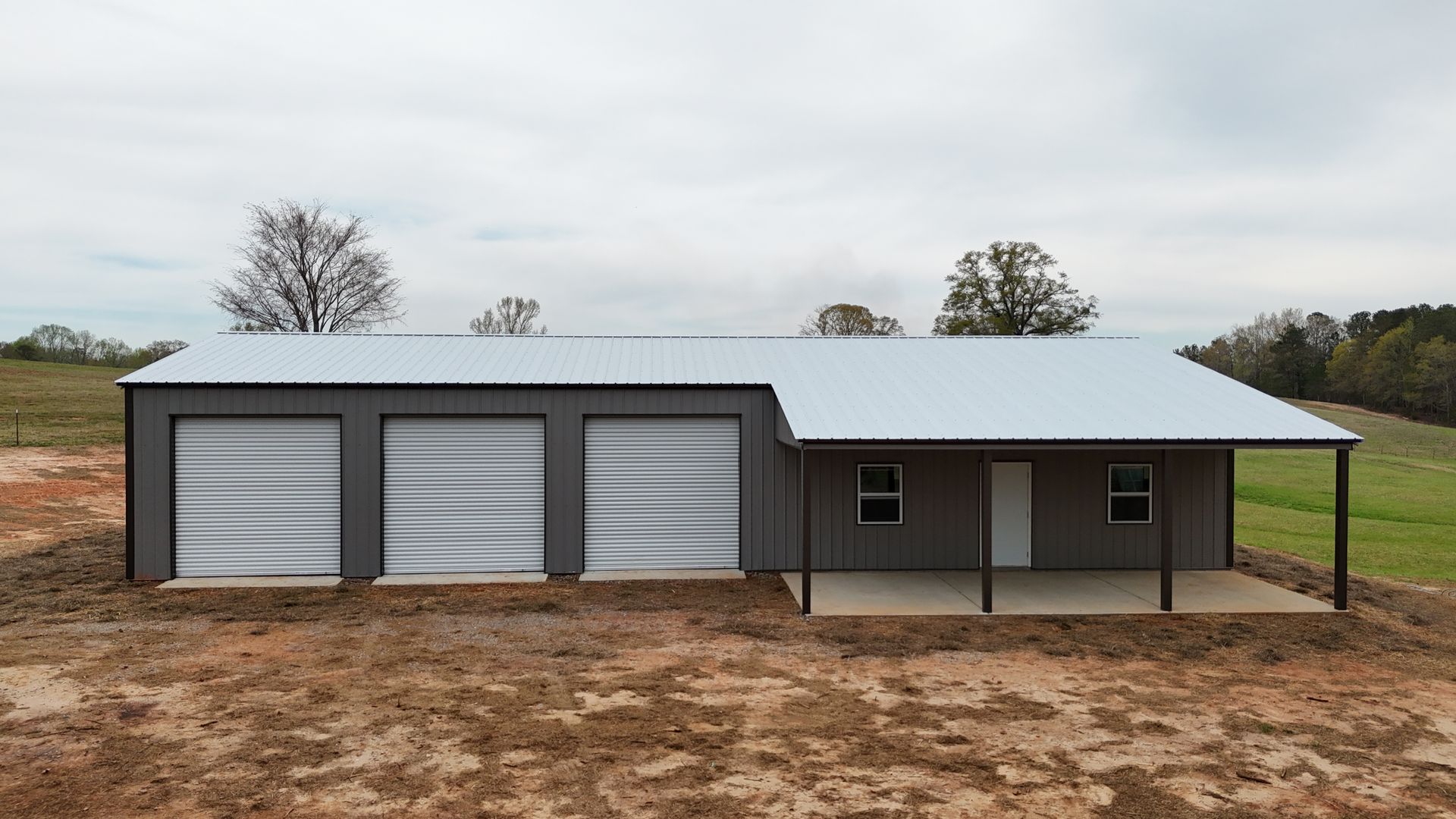 A metal garage with a porch and three garage doors is sitting in the middle of a dirt field.
