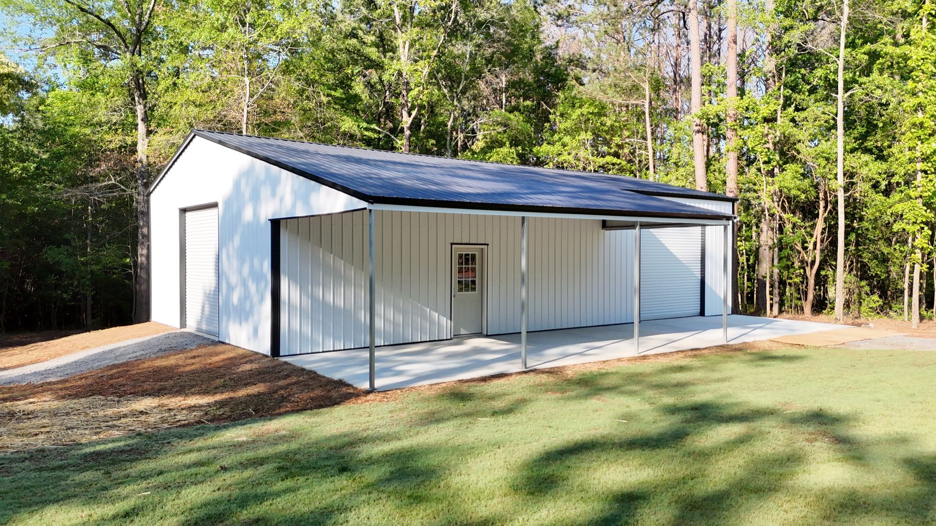 White metal building with black trim, carport, and a concrete pad. Outdoors in a grassy area.