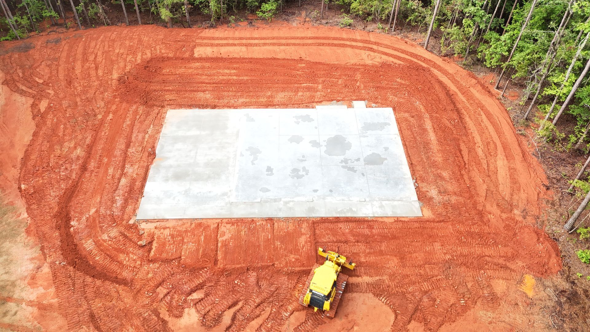 Aerial view of construction site with a concrete foundation surrounded by red soil, and a yellow compact tractor.