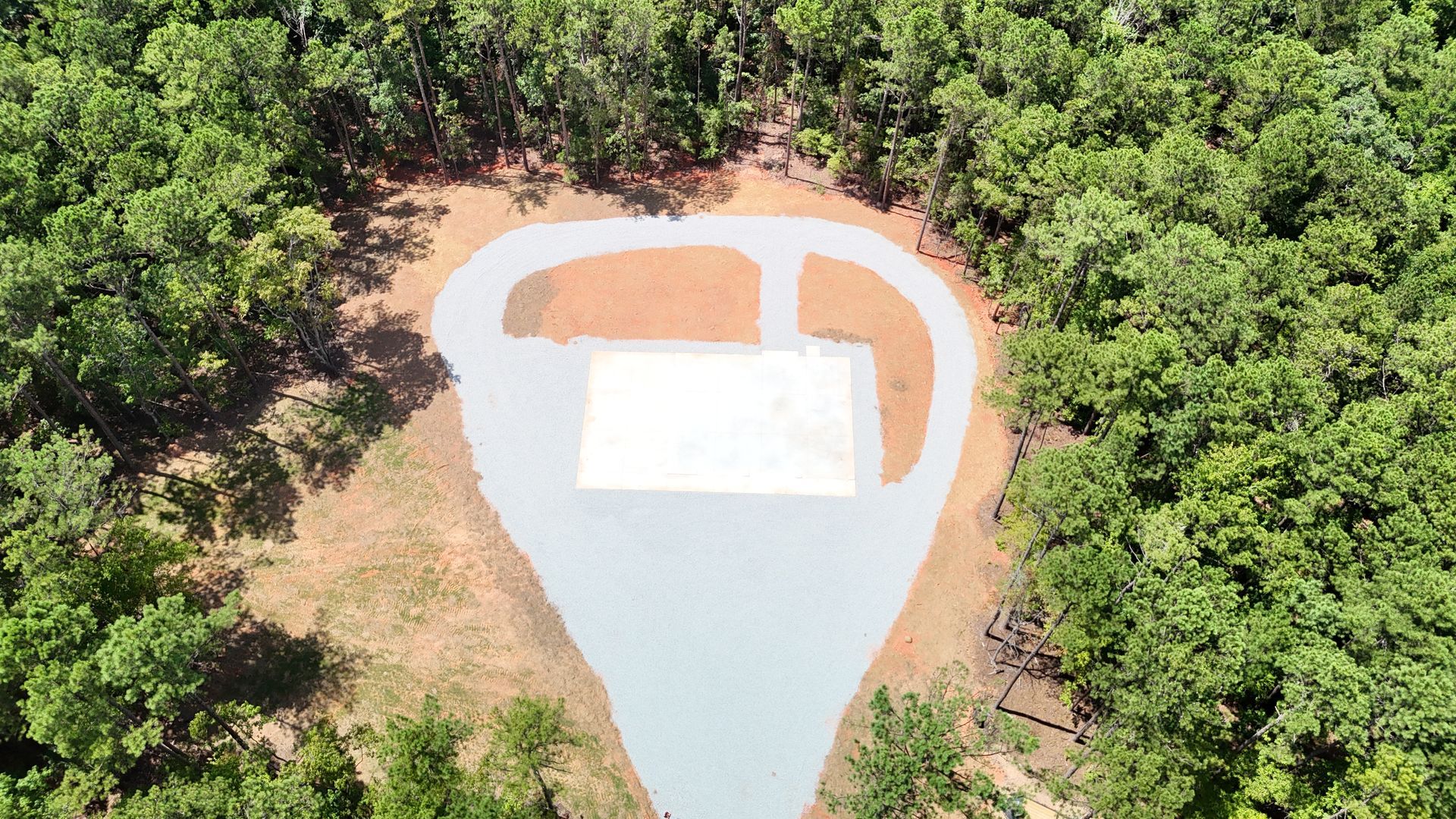 Aerial view of a clearing with a white rectangular structure and paved driveway surrounded by green trees.