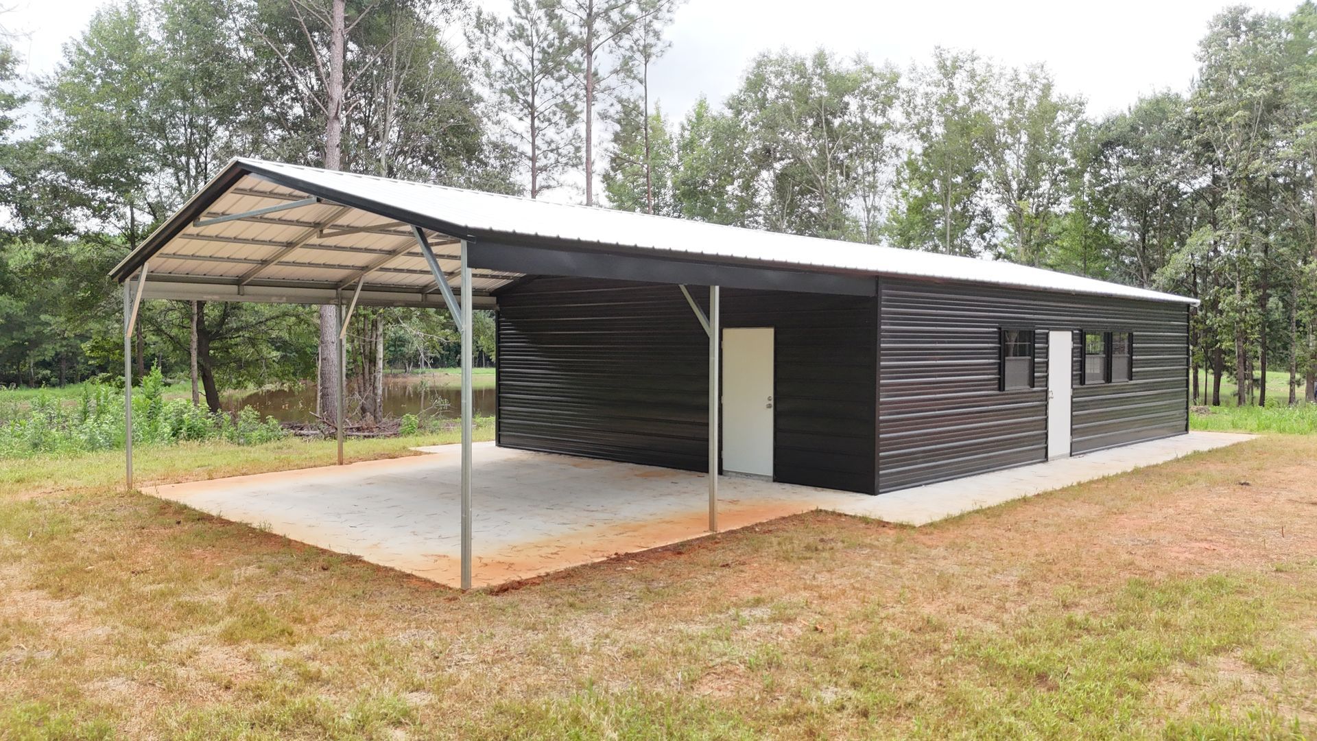 Black metal building with carport, white doors/windows, on a concrete pad, in a grassy area.