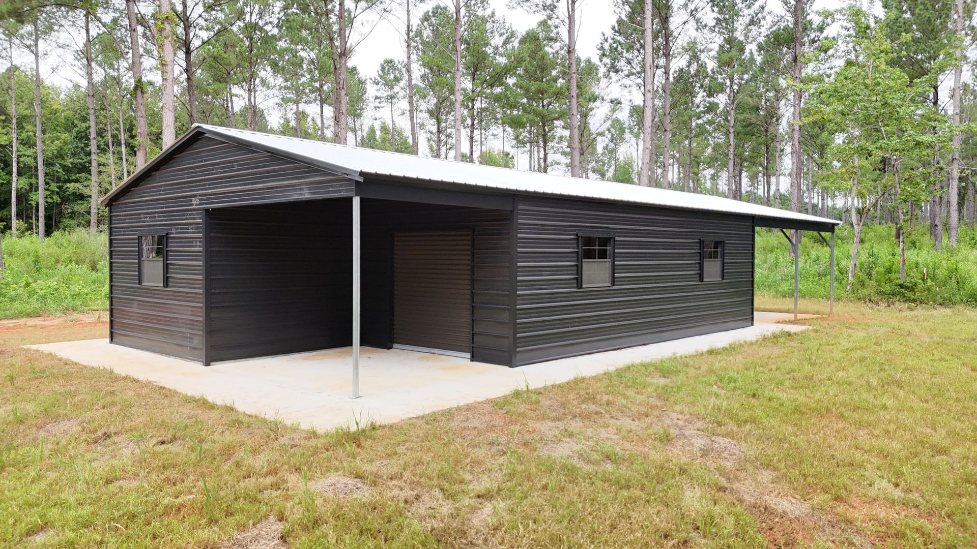 Black shed with a carport, silver roof, on a concrete pad in a grassy area near trees.
