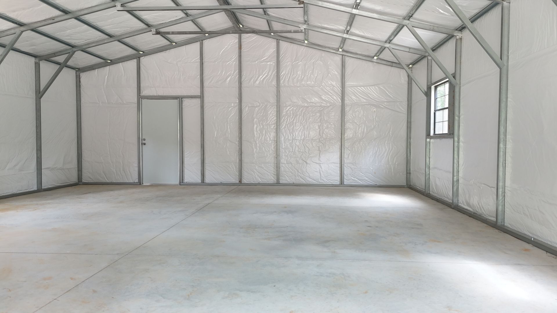 Interior view of an empty metal-framed building with white walls, a concrete floor, and a door and window.
