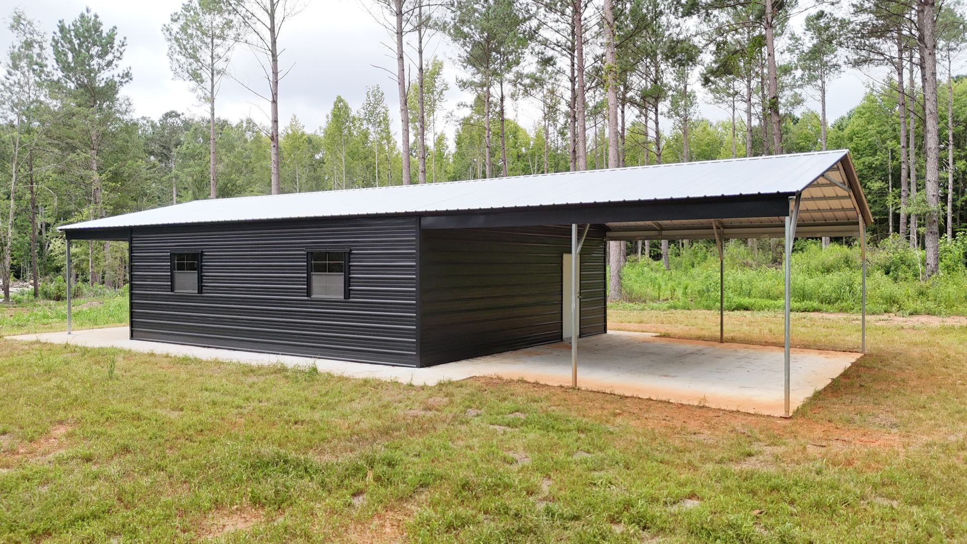 Black metal building with a carport in a grassy area, surrounded by trees.