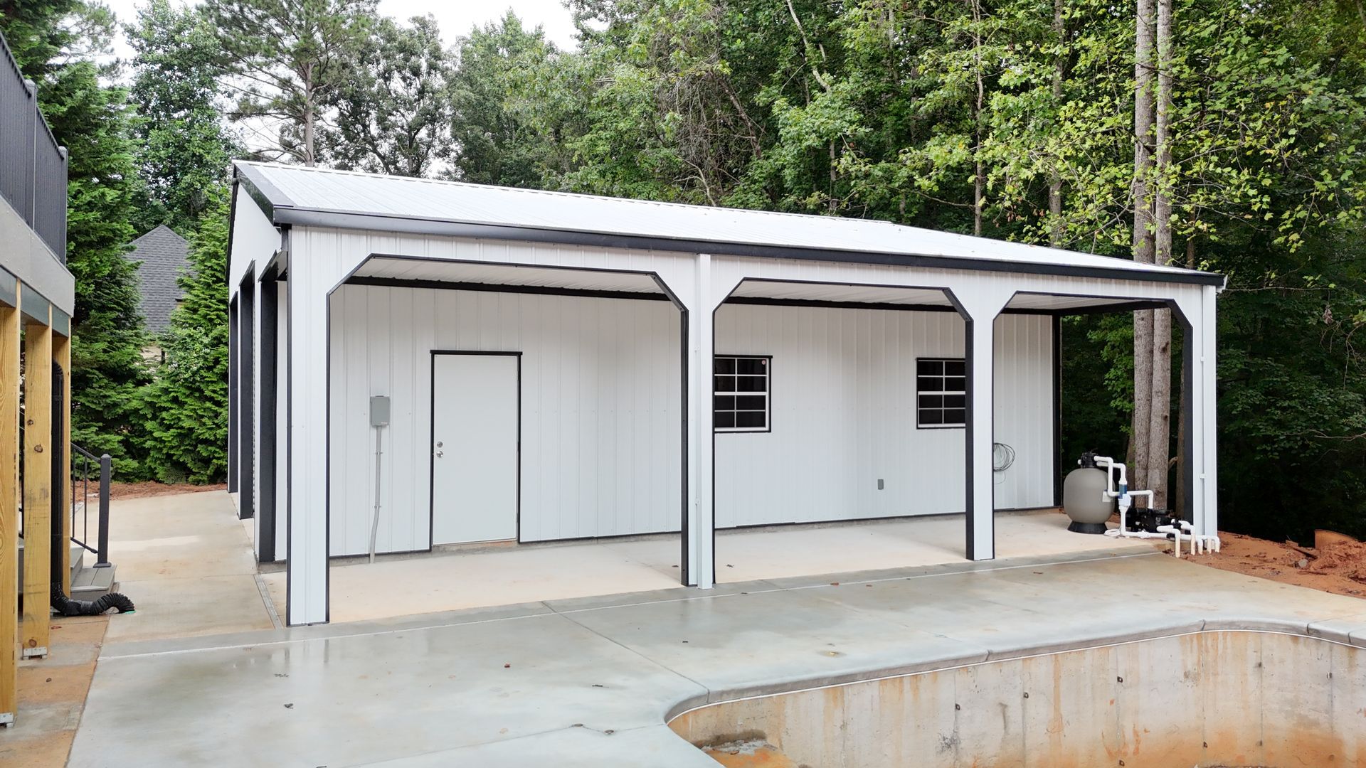 White and black shed with open-air porch; concrete patio in front; adjacent to pool; surrounded by trees.