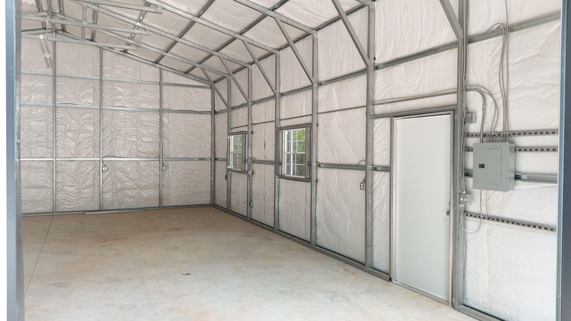 Interior of an unfinished metal building with white insulation, a concrete floor, door, windows, and electrical panel.
