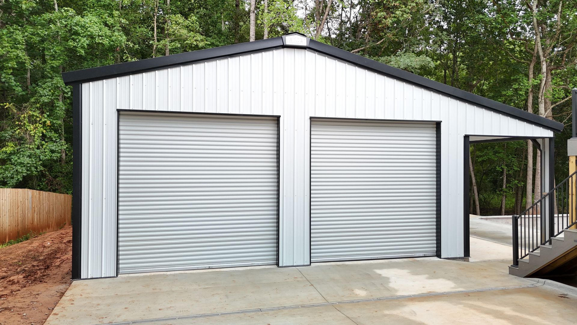 Two-car metal garage with roll-up doors, carport, and black trim. Set on concrete with a wooded backdrop.