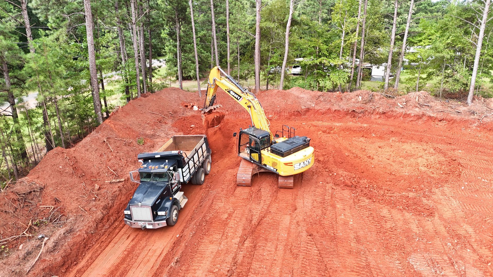 Excavator loading dirt into dump truck on a red-dirt construction site surrounded by trees.