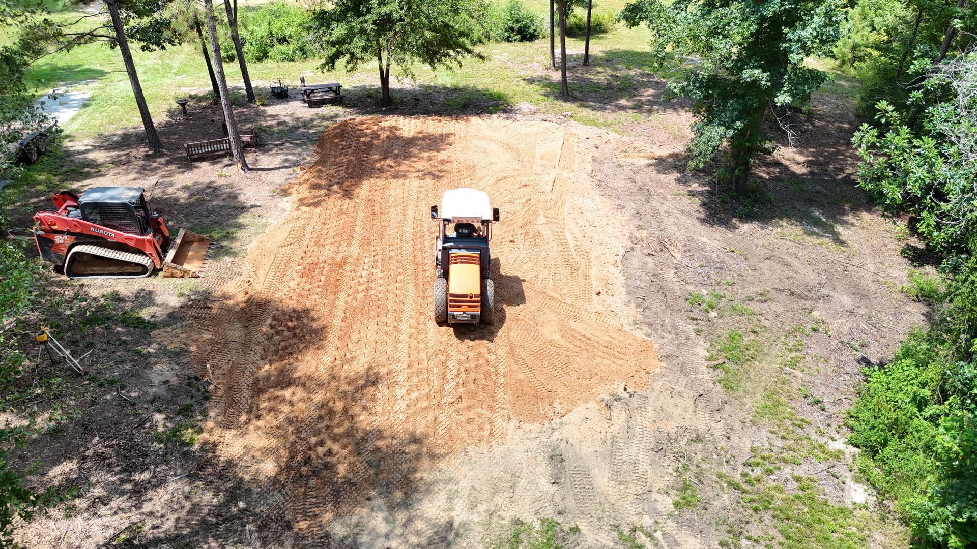 Tractor tilling soil in a clearing, skid steer loader nearby, surrounded by trees.