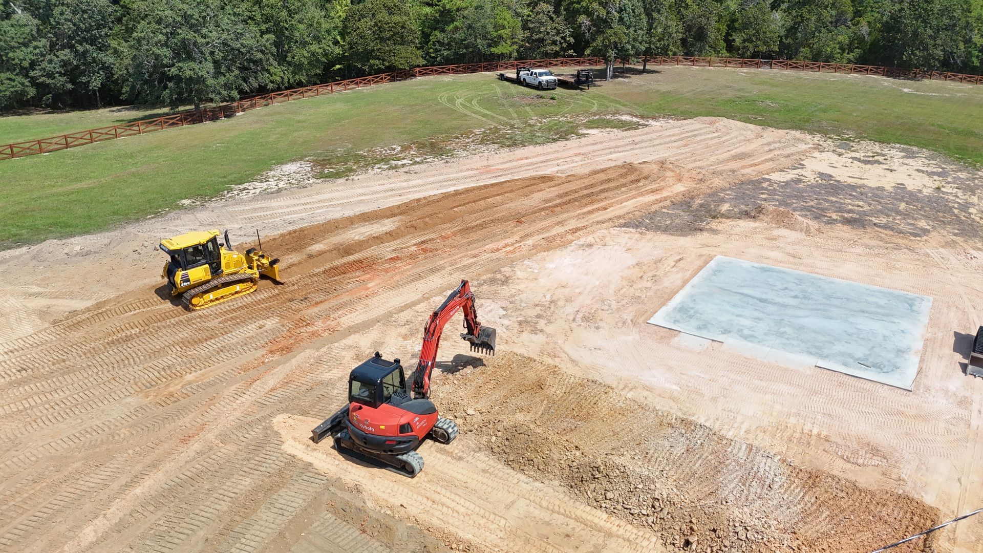 Construction site: bulldozer and excavator working on earth, clearing area near a concrete pad.