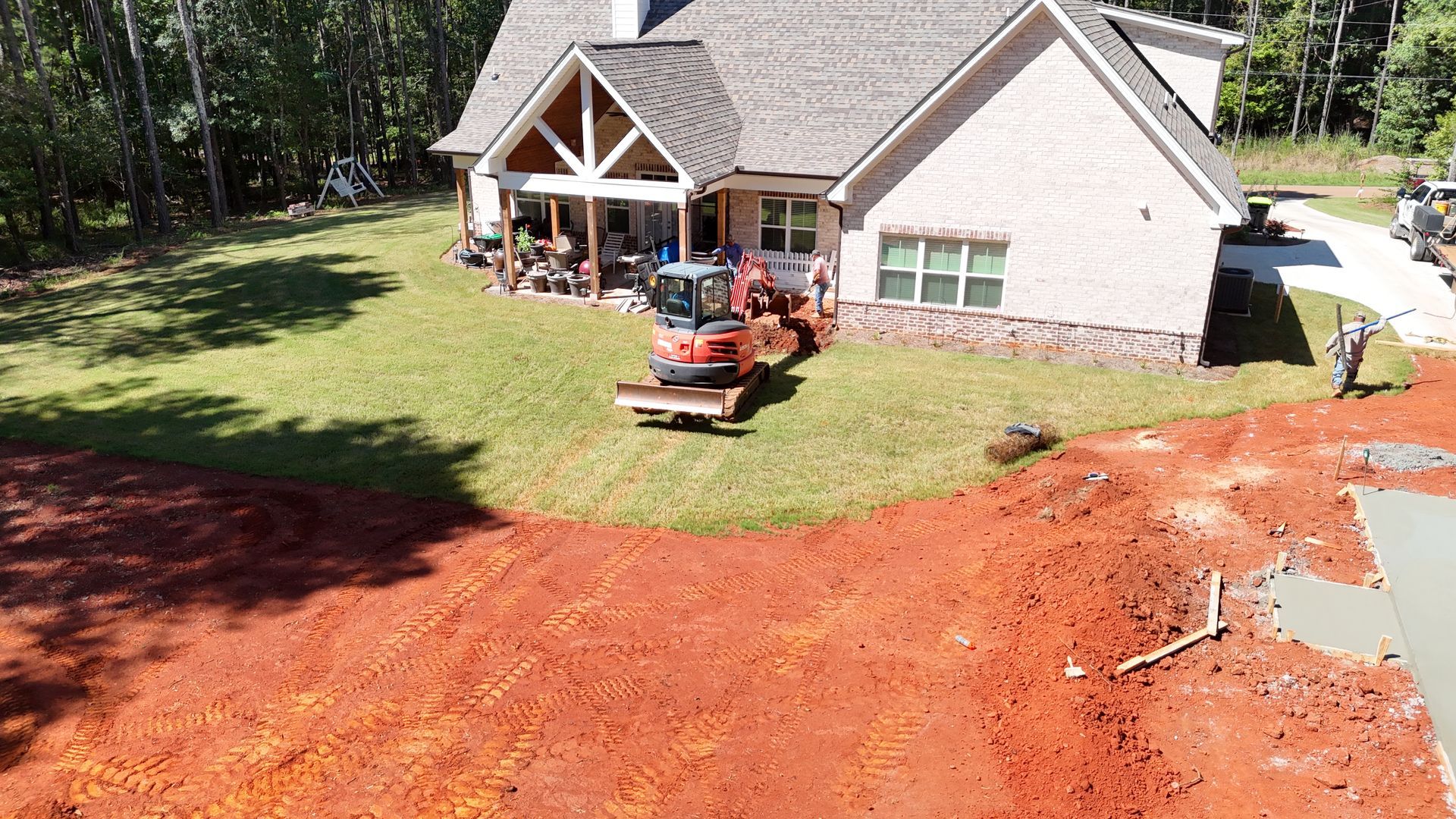 A small tractor grading a lawn in front of a two-story house. Red dirt in the foreground.