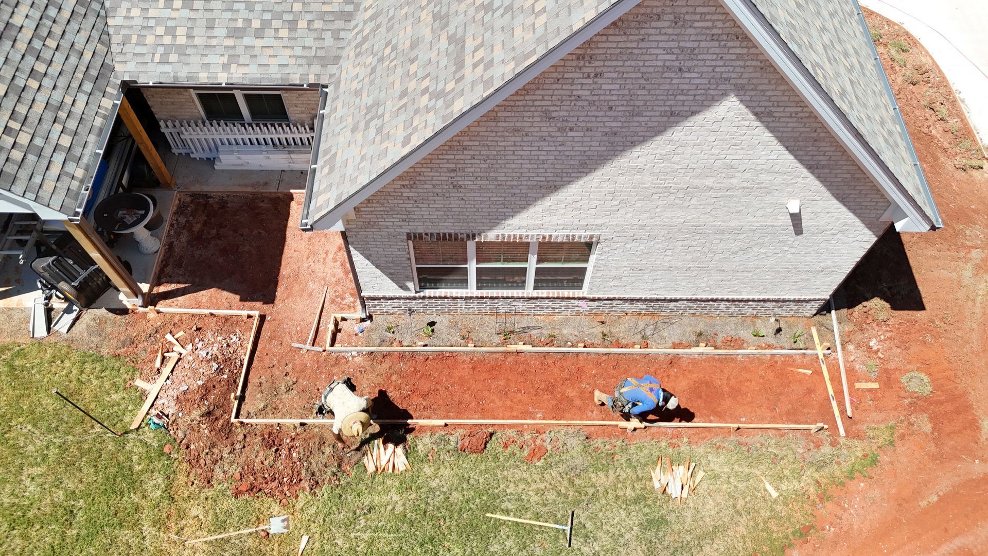Workers laying red brick pathway around a house with gray siding. Green grass surrounds the construction.