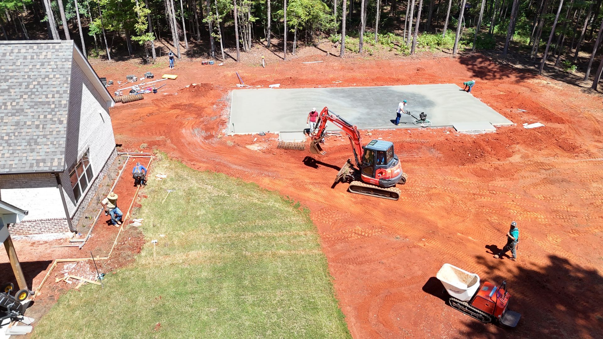 Construction site with excavator, concrete slab, and workers. Red dirt, green grass, house.