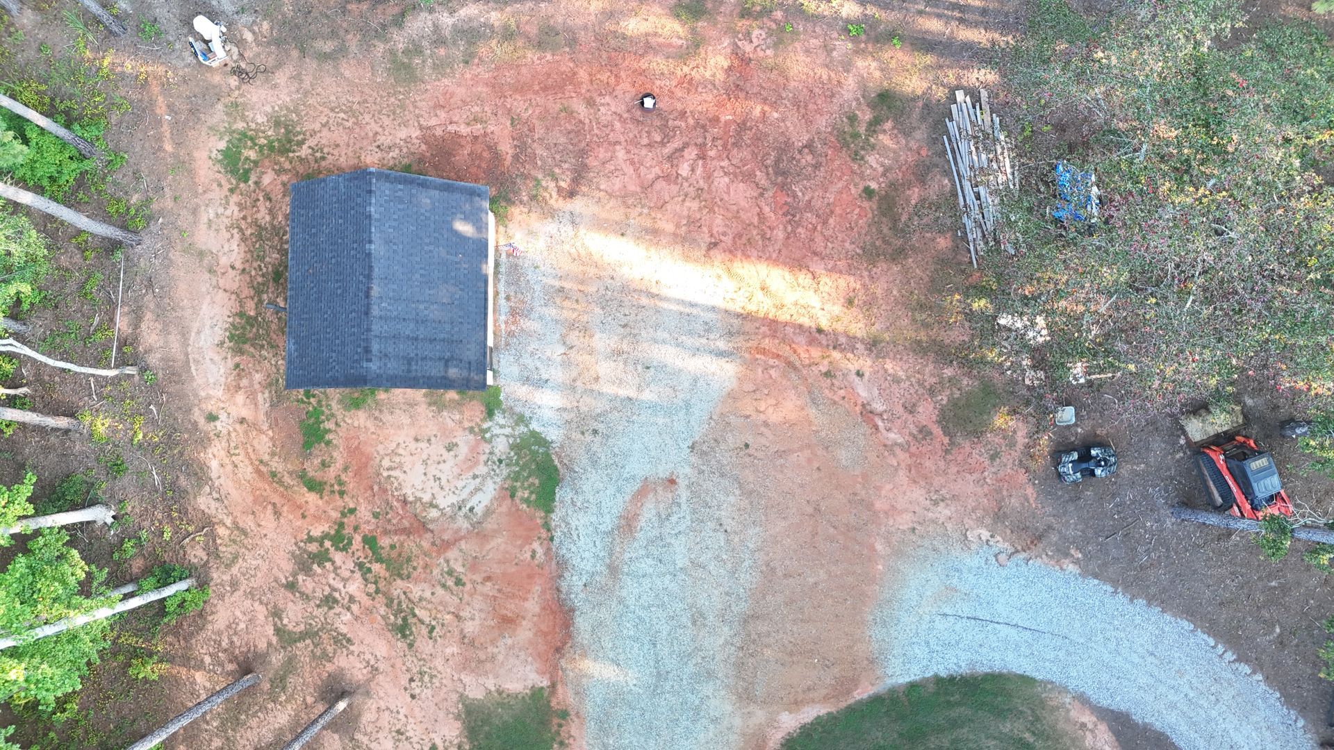 Aerial view of a house under construction in a clearing, with dirt, gravel, and machinery visible.