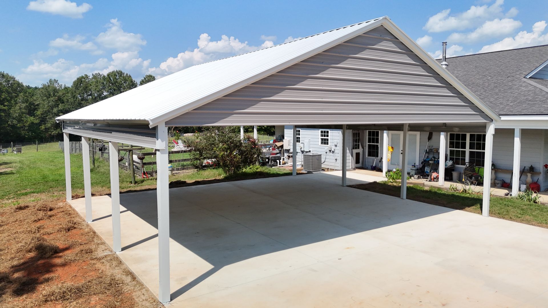 Carport attached to a house; gray and white, concrete pad, blue sky, trees in the background.