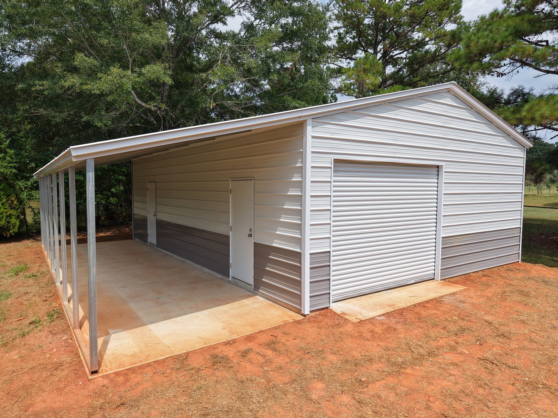Metal garage with carport; white and gray exterior, roll-up door, on dirt ground, trees in background.