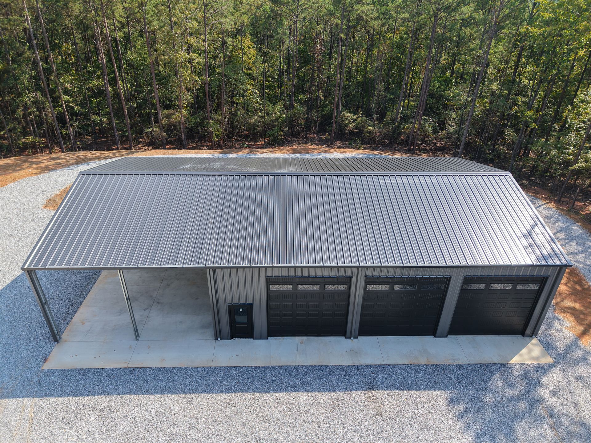 Metal garage building with four garage doors and a carport; gravel driveway and forest background.
