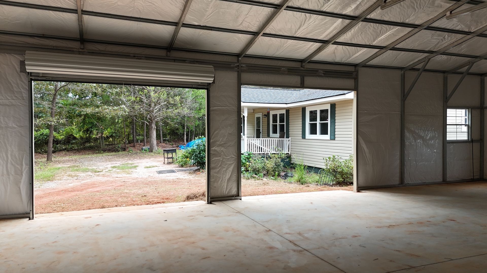 Interior of a gray metal building with open garage doors showing a yard and house.