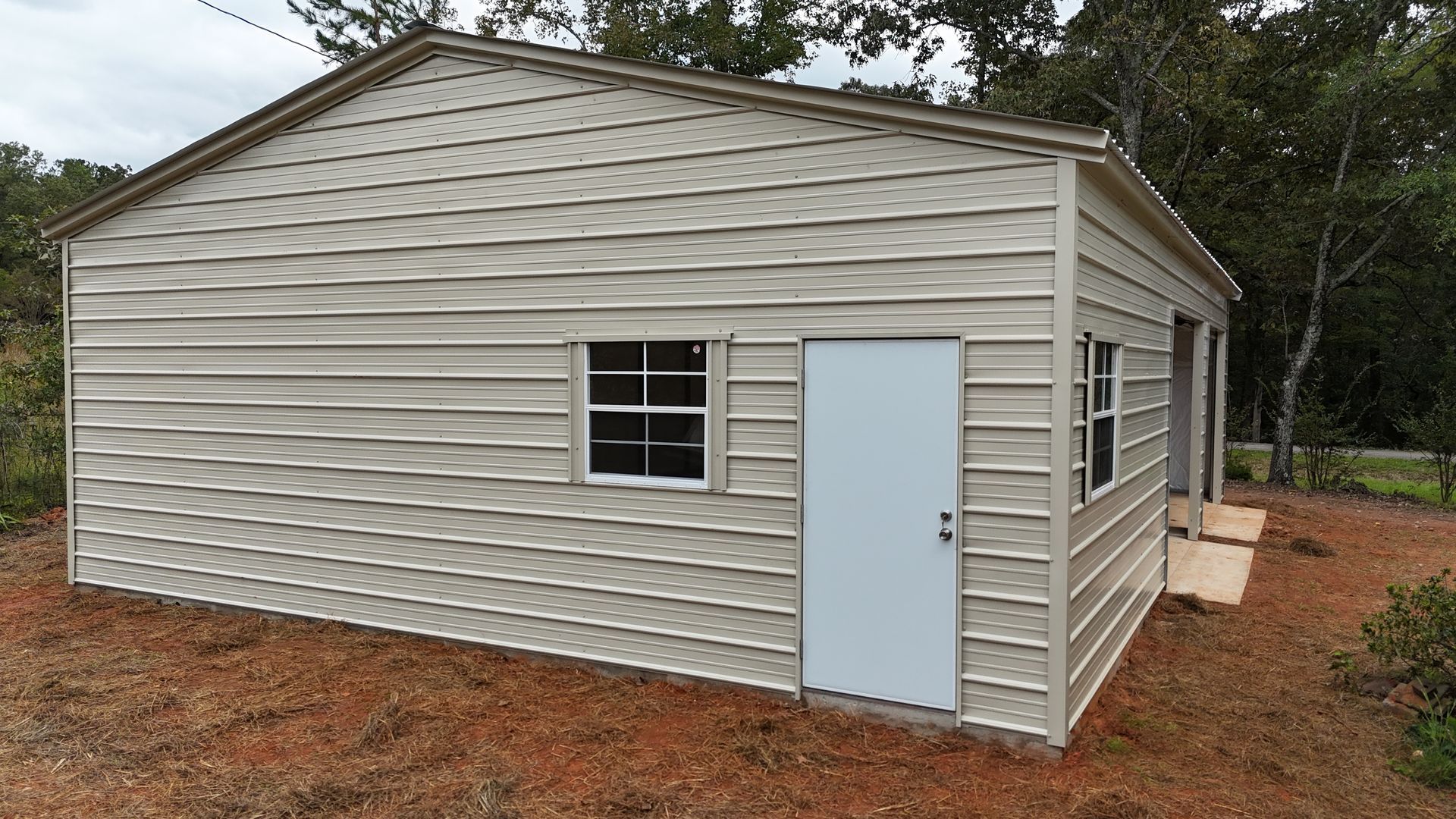 Tan metal shed with a door and two windows on a red dirt lot, surrounded by trees.