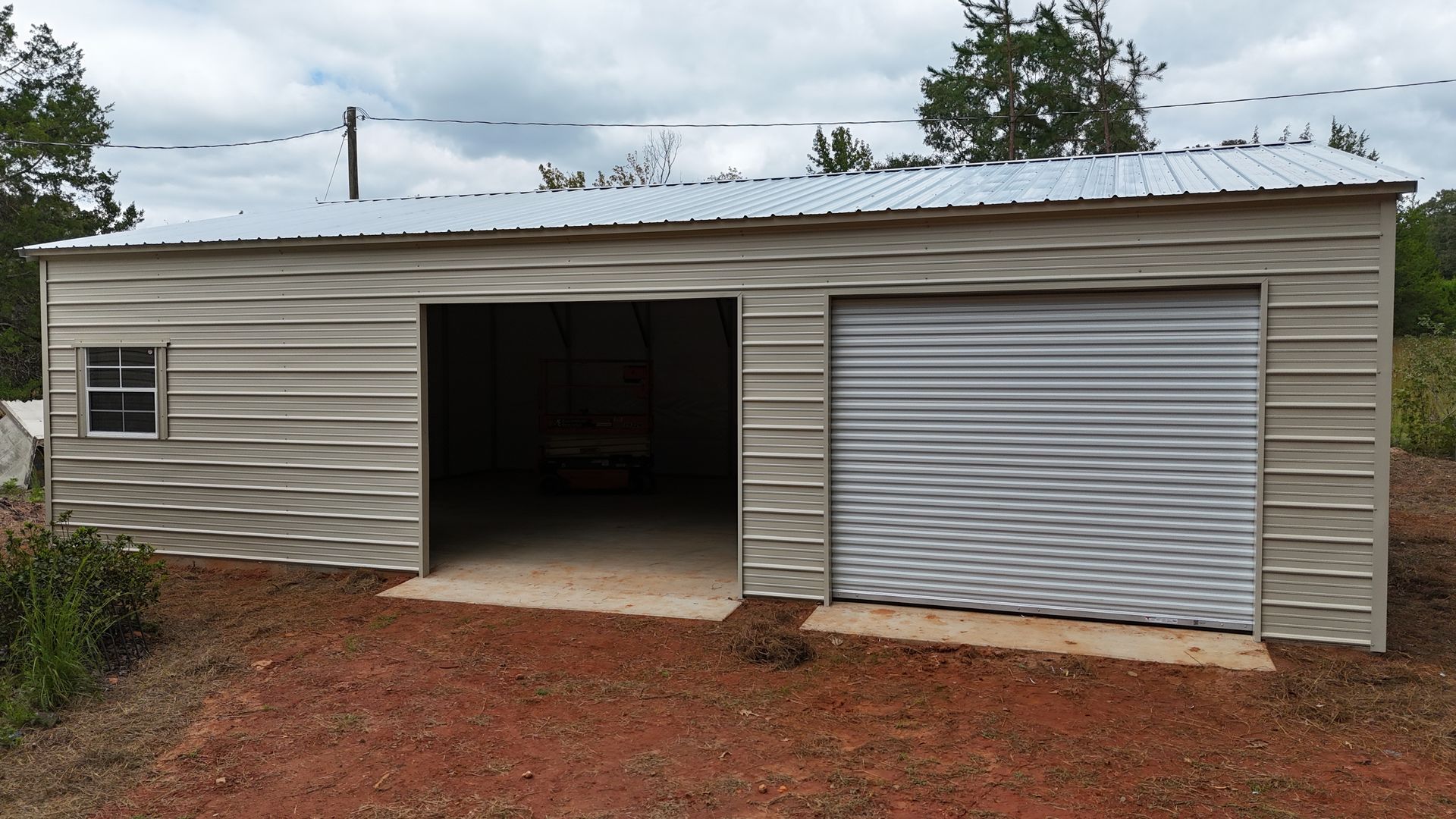 Tan metal garage with open bay, closed roll-up door, and small window, on red dirt, under a cloudy sky.