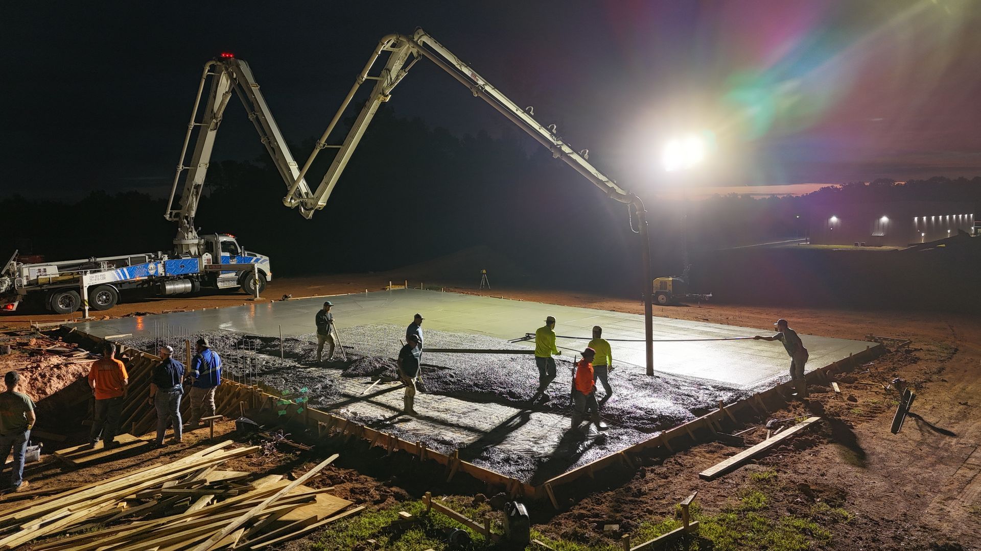 Construction workers pouring concrete at night with a large pump truck and bright lights illuminating the work site.