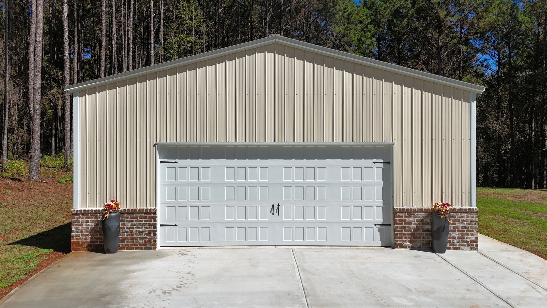 Beige metal garage with white garage door, brick base, and concrete driveway, set against trees.
