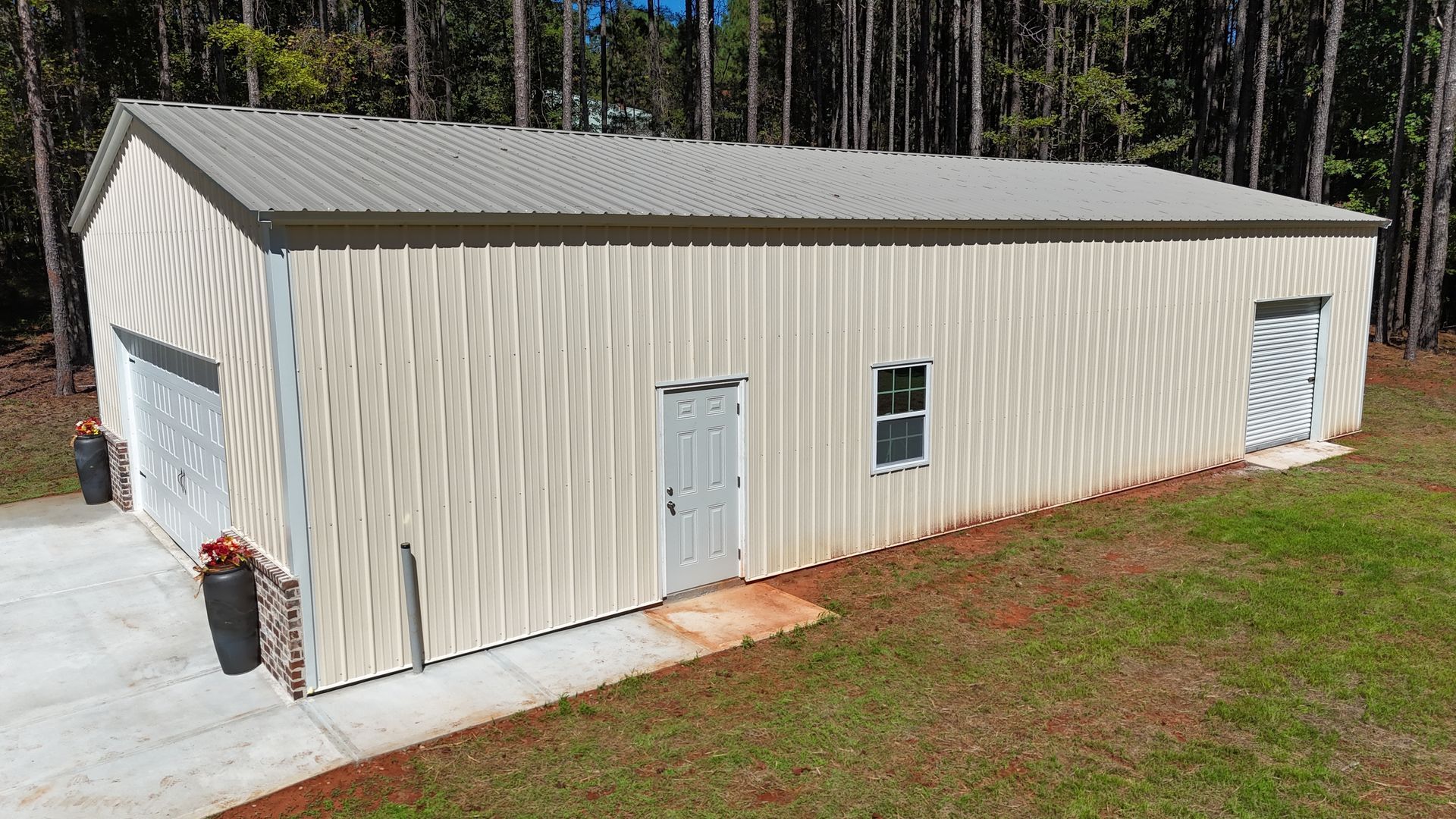 Beige metal building with garage doors, door, and window on green grass near trees.
